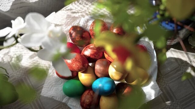 A vibrant collection of Easter eggs rests on a table under soft, natural sunlight, casting delicate shadows. The close-up shot highlights the rich colors and textures.