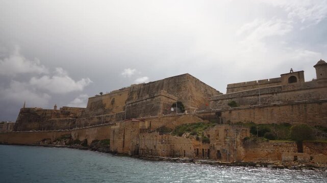 View from passing boat along coastal walls of Fort St Elmo in Valletta. Overcast but warm weather. Massive limestone fortification rises above sea, showcasing its historic military architecture