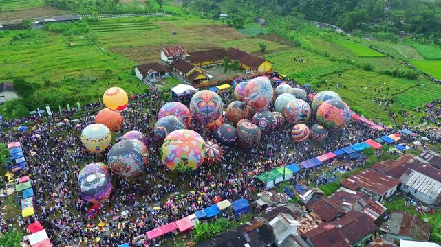 Bright balloons rising over green fields during a festival. Cinematic drone view with crowd, tents, and scenic hills. Wonosobo balloon festival, Indonesia.