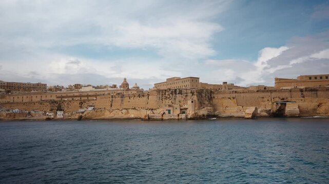 View from moving boat of coastal walls and structures of Fort St Elmo in Valletta. Massive limestone fortifications line waterfront under soft cloudy light, highlighting historic military architecture