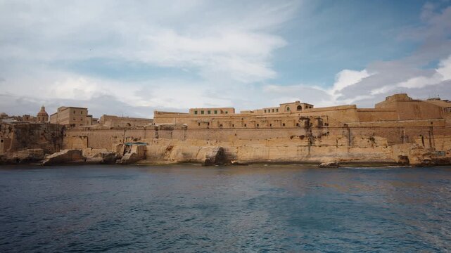 View from moving boat of coastal walls and structures of Fort St Elmo in Valletta. Massive limestone fortifications line waterfront under soft cloudy light, highlighting historic military architecture