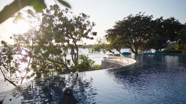 A tranquil pool by the beach during sunrise with warm light reflecting on the water. Green trees frame the scene while the ocean and shoreline create a peaceful tropical coastal atmosphere.