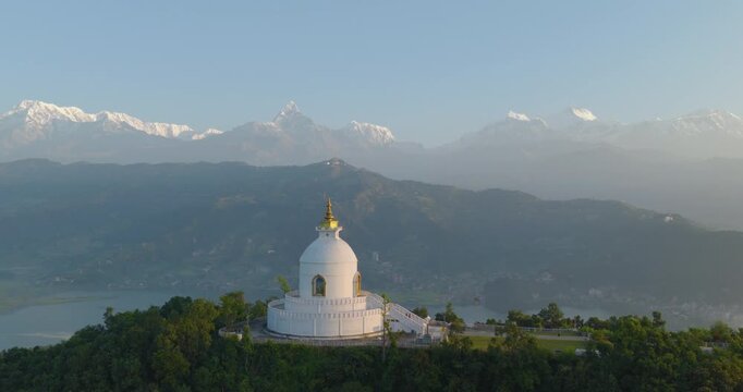 Drone pull in shot approaching the Buddhist monument World Peace Stupa in Pokhara, Nepal, with lush green forest and Annapurna and Machhapuchhre peaks in the background.