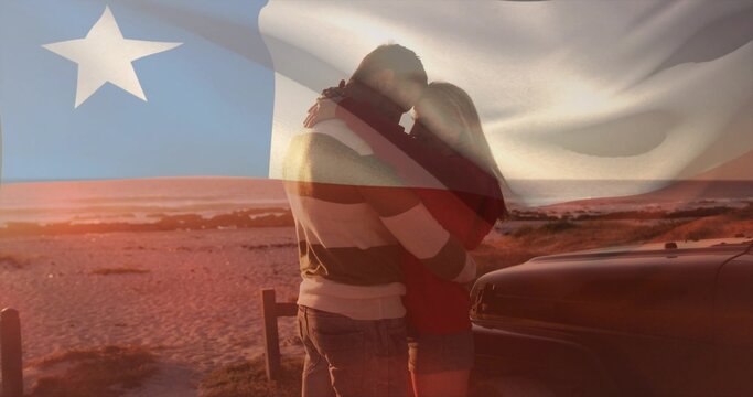 Embracing couple wearing red top, shorts on beach at sunset, beside car, waves, flag overlay
