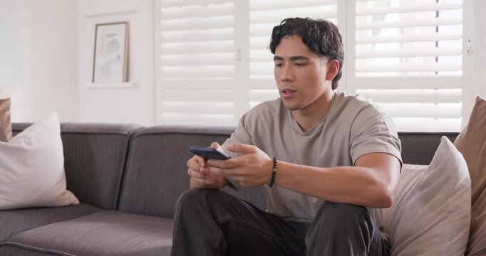 Asian man glancing right, reaching for phone on sofa in bright living room, using voice command