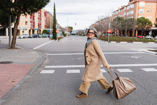 Woman walking joyfully across city street in winter coat with bag