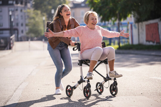 Smiling granddaughter pushes grandmother with walker outdoors in city