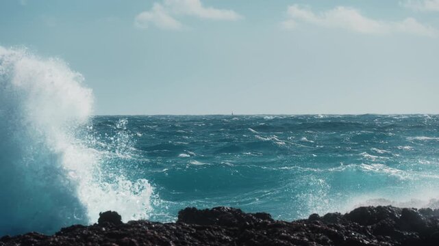Slow motion waves shimmer in bright sunlight as wind drives them across open sea to the horizon, crashing near a rocky shoreline in foreground. Clean seascape with vivid water color and dynamic motion
