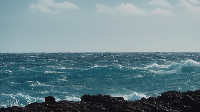 Slow motion waves shimmer in bright sunlight as wind drives them across open sea to the horizon, crashing near a rocky shoreline in foreground. Clean seascape with vivid water color and dynamic motion