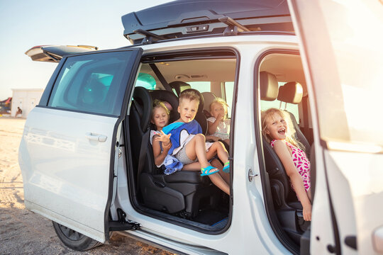 Group of four cute adorable little happy caucasian children enjoy having fun sit in minivan going to sea beach road trip on hot summer day. Kids in swimsuit in car with side open door against sun