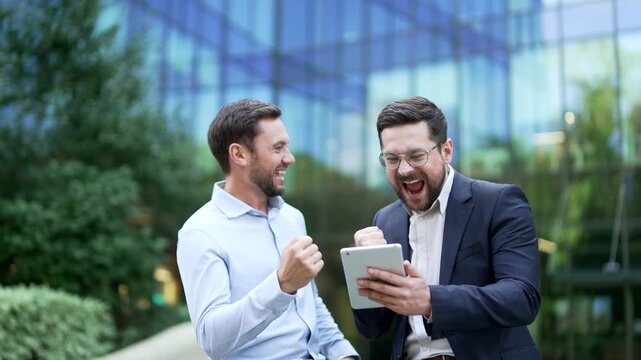 Two happy excited businessmen standing outdoors using a digital tablet, reacting to successful online results and a closed business deal, celebrating profit, achievement, and growth, showing teamwork