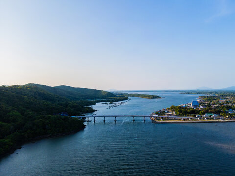 Aerial view of calm river winding through green hills. Bridge spans water, connecting land masses. Town with buildings lines the riverbank. Sky is clear, sunlight glows gently over hills