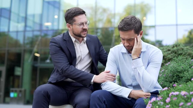 Coworker comforting a stressed businessman sitting on a bench outdoors near office building, offering support and understanding, showing care, empathy, emotional connection in difficult work situation