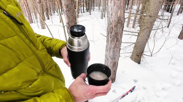 Hiker In Woods Brews Warm Beverage Amid Snow. Winter Hiker Pauses To Enjoy Hot Drink Beside Skis. Man In Green Jacket Takes Break With Steaming Coffee Under Pine Trees During Winter Hike