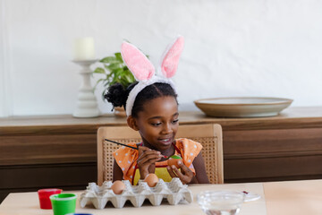 Naklejka premium African American female child painting Easter egg at wooden table holding paintbrush and egg