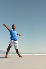 African American man standing barefoot on sandy beach at waterline wearing blue shirt white shorts © wavebreak3