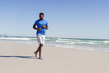 African American man jogging along sandy shore, in blue-tee-shoes-watch, waves-point, copy space © wavebreak3