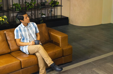 Asian man sitting on leather sofa in lounge with potted plants holding dark mug, looking up-left © wavebreak3