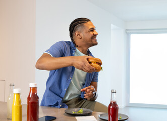 Man in blue shirt holding hamburger at kitchen counter, responding to offframe voice, offering bite © wavebreak3