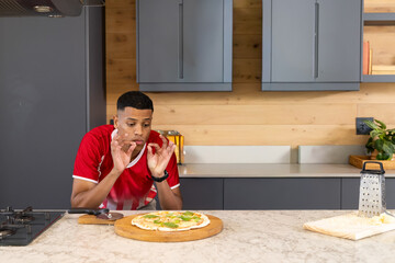 African American man seasoning pizza at counter in home kitchen, pizza board, grater, in red shirt © wavebreak3
