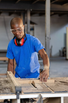 African American man operating table saw, guiding plank in shop with blue tee, goggles and earmuffs