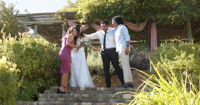 Wedding party huddling under pergola, linking and guiding bride in gown holding bouquet down steps