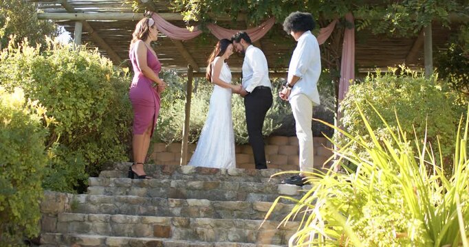 Couple in wedding attire holding hands, leaning heads during vows on stone steps beneath pergola