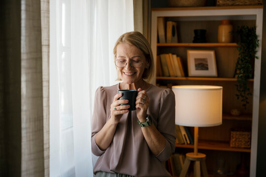Smiling woman with cup standing by window in cozy home interior