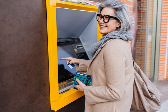 Woman withdrawing cash at outdoors ATM wearing scarf and glasses