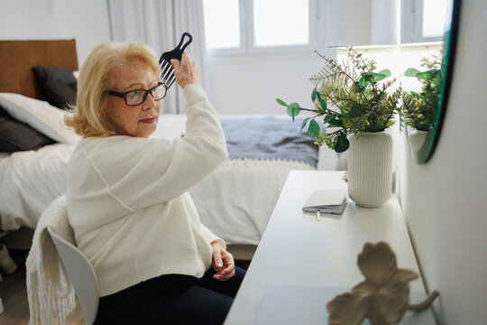Woman combing hair in bedroom sitting at desk by mirror