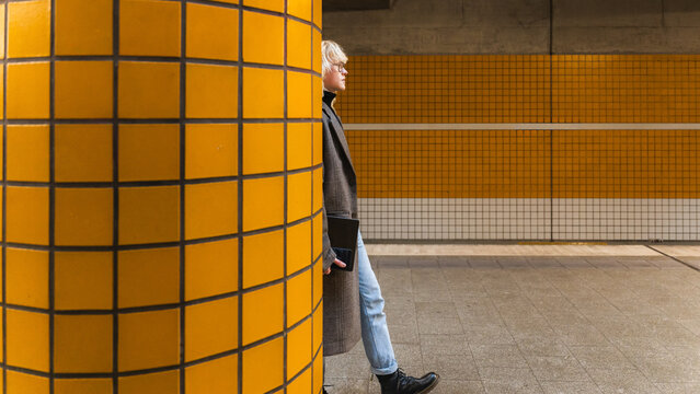 Young mann waiting at subway station with smartphone and leaning on yellow tiled pillar
