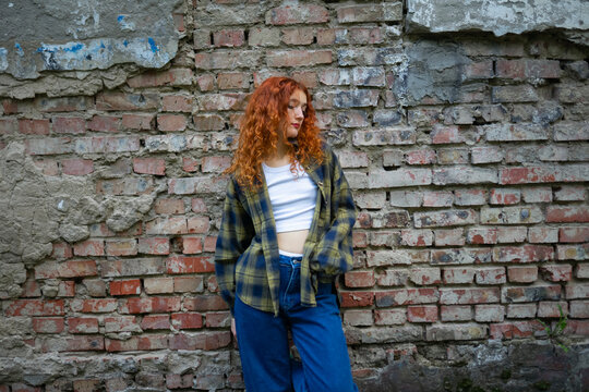 Red-haired woman in casual outfit posing by old brick wall
