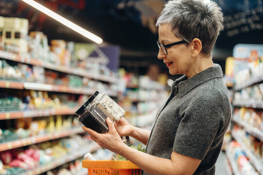 Mature woman with short gray hair examines spice jars while shopping in a grocery store aisle, colorful products displayed on shelves in the background