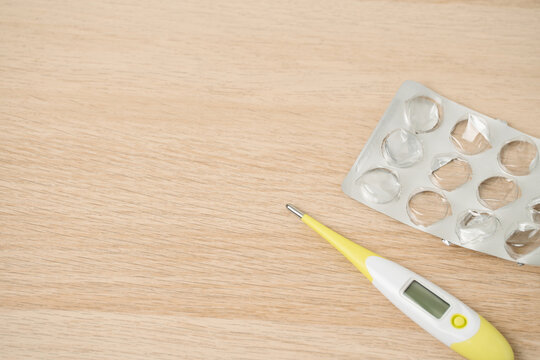 A digital thermometer lies next to an empty blister pack on a light wooden table, suggesting fever, medication use and home healthcare routines focused on recovery and everyday medical essentials.