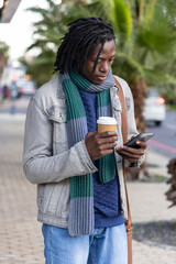 African American man standing on sidewalk checking phone holding coffee wearing scarf © wavebreak3