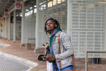 Mid adult African American man waiting at bus stop wearing green scarf carrying bag holding phone © wavebreak3