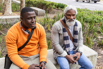 African American men sitting on concrete seats at roadside holding smartphone wearing denim jacket © wavebreak3