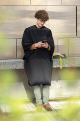 Male graduate wearing gown leaning on stone ledge in courtyard holding smartphone, cap and diploma © wavebreak3