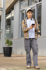 Asian man standing on sidewalk by windows wearing plaid shirt using phone with backpack © wavebreak3