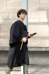 Male graduate wearing black gown walking on plaza holding cap with yellow tassel using smartphone © wavebreak3