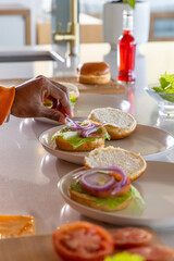 African American man placing red onion rings on bun at kitchen countertop with lettuce tomato © wavebreak3