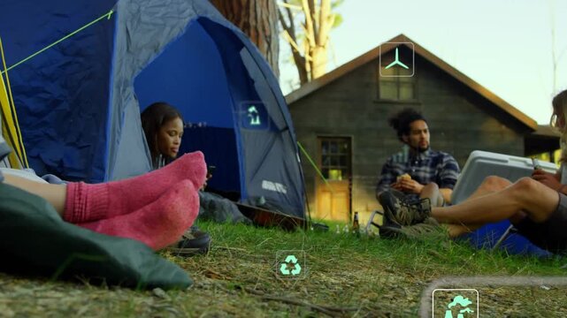 Diverse friends sitting at grassy campsite by cabin monitoring AR data near blue tent