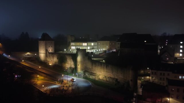 Night drone shot revealing Tour Jacob in Luxembourg, surrounded by illuminated buildings and city lights, capturing the historic atmosphere and vibrant urban skyline.