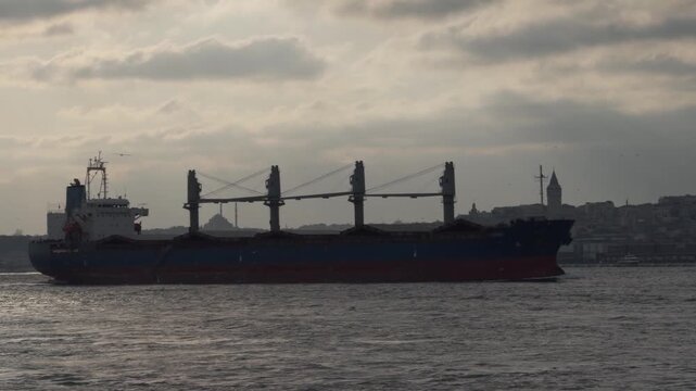 A large logistics ship passing through the Bosphorus, with dense buildings, hotels, and city skyline on the European side. Maritime transport and urban life together.