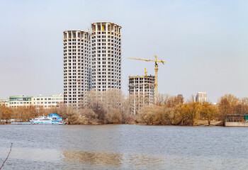 Obraz premium Scenic spring landscape showing city encroaching on nature with tall residential buildings under construction on a river bank. Concrete skyscrapers contrast with trees and water on a sunny April day. 