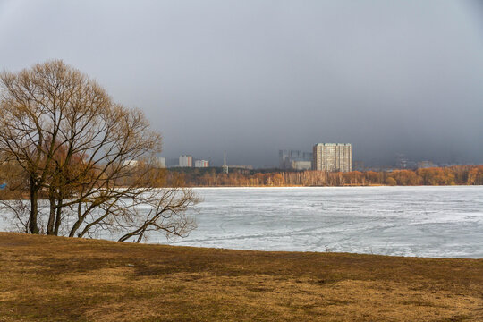 Scenic spring landscape in Strogino with Moskva River floodplain covered in loose melting ice. Bare trees on a muddy shore under a dark cloudy sky with urban buildings in the distance. Russia.