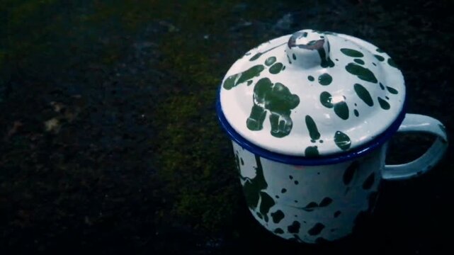 Old school style, a green white abstract pattern mug made of zinc and coated with enamel usually used for drinking hot tea or coffee under rain drop water, with grunge mossy floor dark background. 
