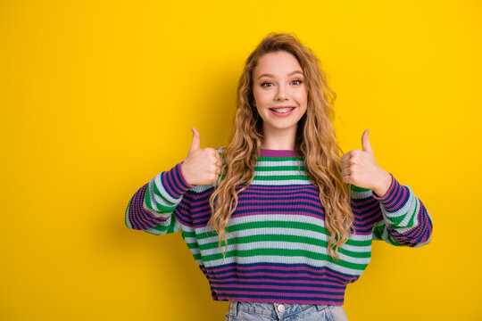Young female model with striped pullover sweater smiles and shows thumbs up against bright yellow background