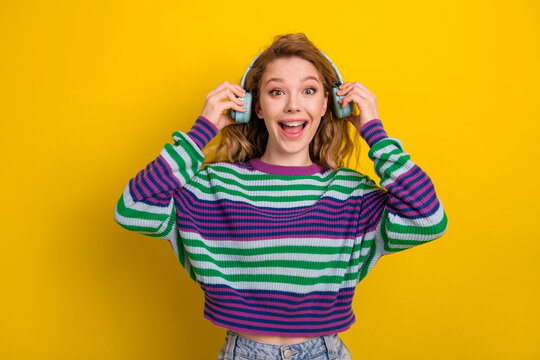 Young woman wearing striped sweater listening to headphones smiling against a bright yellow background