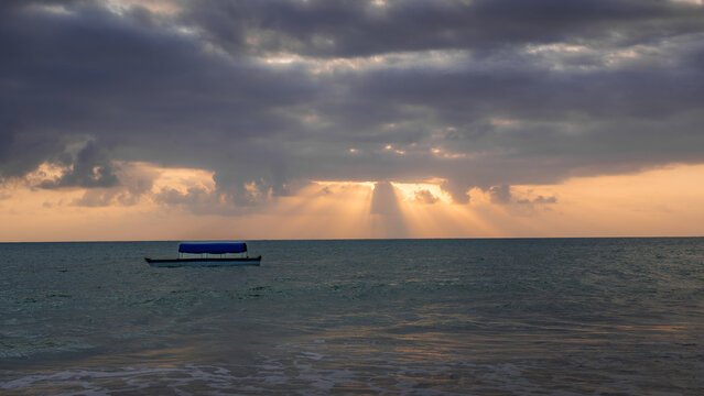 View of the sun's rays pierce through the dark, dramatic clouds, illuminating the tranquil turquoise ocean and a small boat, Paje, Tanzania.
